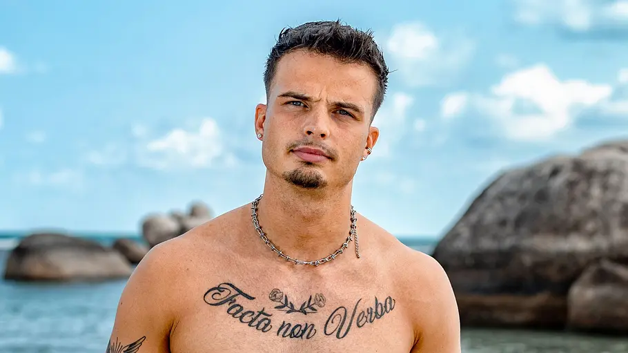 Brian Ein tätowierter Mann steht oberkörperfrei am Strand und blickt mit ernstem Ausdruck in die Kamera. Er trägt rote Badehose und Schmuck, während hinter ihm das Meer, große Felsen und ein leicht bewölkter Himmel zu sehen sind.