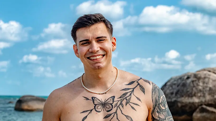 Cansin Ein tätowierter Mann steht oberkörperfrei am Strand und lächelt in die Kamera. Er trägt eine auffällige neongelbe Badehose und Schmuck, während im Hintergrund das Meer, Felsen und ein leicht bewölkter Himmel zu sehen sind.