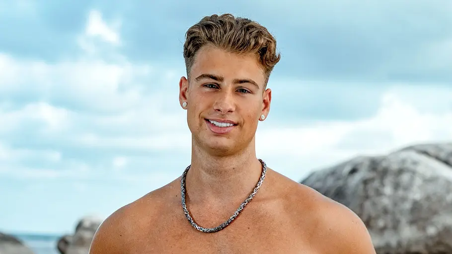 Daymian Weiß Ein junger Mann steht oberkörperfrei am Strand und lächelt in die Kamera. Er trägt eine rosafarbene Badehose und Schmuck, während im Hintergrund das Meer, große Felsen und ein leicht bewölkter Himmel zu sehen sind.