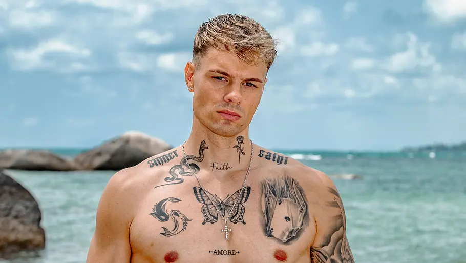 Fabi Hesdahl Ein tätowierter Mann steht oberkörperfrei am Strand und blickt ernst in die Kamera. Er trägt eine orangefarbene Badehose, während hinter ihm das Meer, Felsen und ein leicht bewölkter Himmel zu sehen sind.