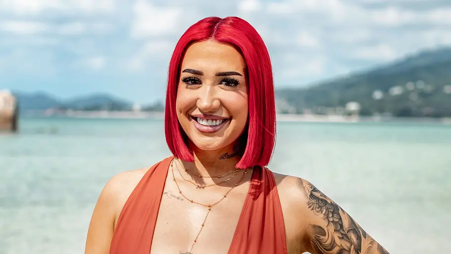 Francesca Brinley Eine Frau mit auffällig roten Haaren steht lächelnd am Strand und blickt in die Kamera. Sie trägt einen einteiligen Badeanzug, Schmuck und hat mehrere Tattoos, während im Hintergrund das Meer, Felsen und eine hügelige Küstenlandschaft zu sehen sind.