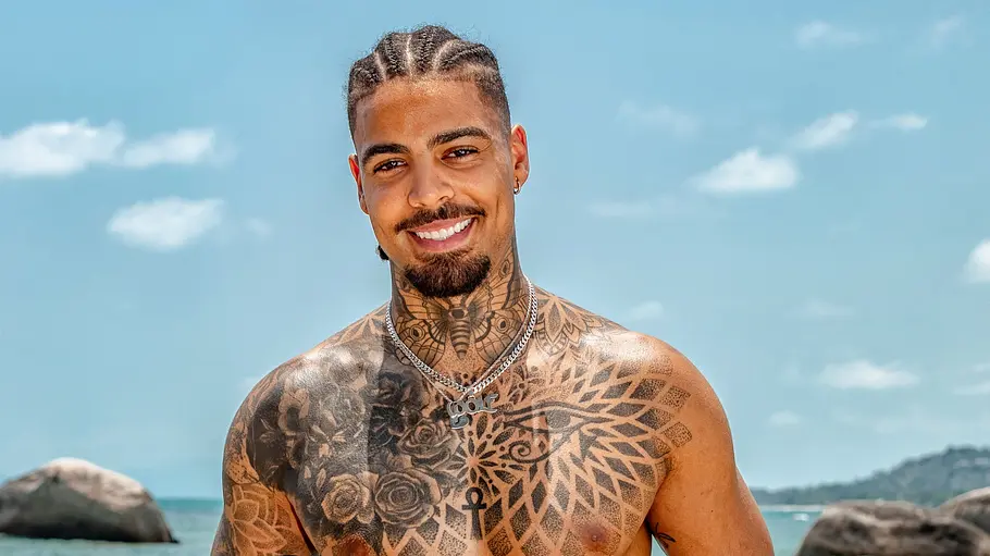 Germain Wolf Ein stark tätowierter Mann steht oberkörperfrei am Strand und lächelt in die Kamera. Er trägt eine Badehose und Schmuck, während hinter ihm das Meer, Felsen und eine sonnige Küstenlandschaft zu sehen sind.