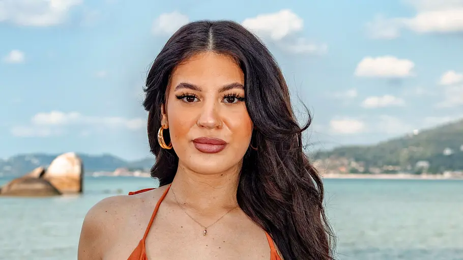Janice Eine Frau im orangefarbenen Bikini steht am Strand und blickt direkt in die Kamera. Sie hat lange dunkle Haare, trägt Schmuck und hat mehrere Tattoos, während im Hintergrund das Meer, Felsen und eine hügelige Küstenlandschaft zu sehen sind.