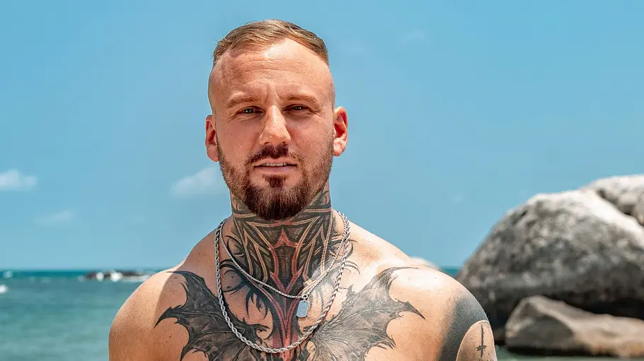 Johannes Ein tätowierter Mann steht oberkörperfrei am Strand und blickt direkt in die Kamera. Er trägt eine gemusterte Badehose und Schmuck, während im Hintergrund das Meer, große Felsen und ein sonniger Himmel zu sehen sind.