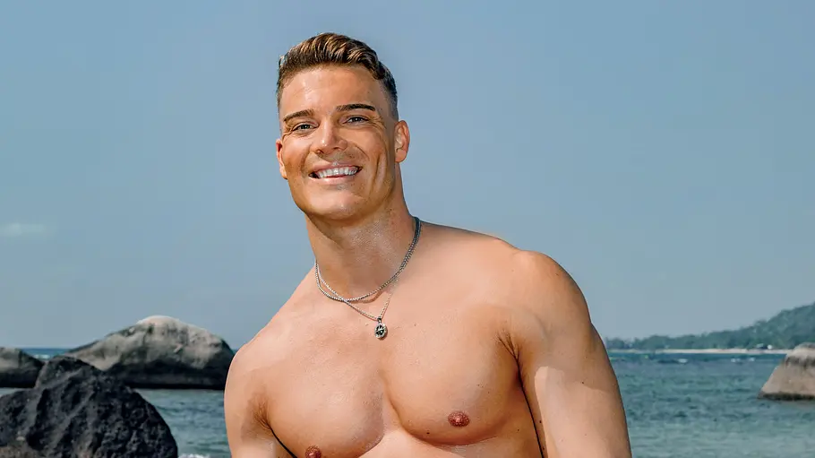 Marwin Klute Ein muskulöser Mann steht oberkörperfrei am Strand und lächelt in die Kamera. Er trägt eine rosafarbene Badehose und Schmuck, während im Hintergrund Felsen, Meer und eine ruhige Küstenlandschaft zu sehen sind.