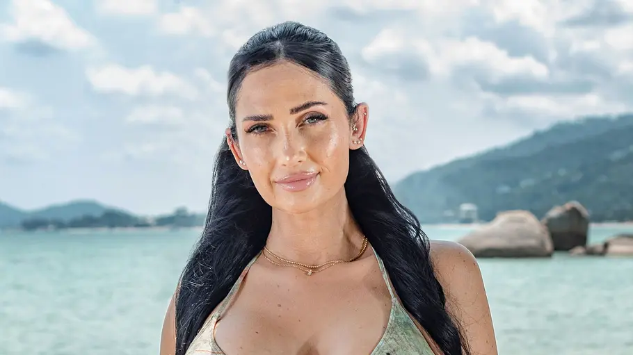 Michelle Bendig Eine Frau im Bikini steht am Strand und blickt mit leichtem Lächeln in die Kamera. Sie trägt Schmuck, hat lange dunkle Haare und wird von einer Küstenlandschaft mit Meer, Felsen und wolkigem Himmel im Hintergrund eingerahmt.