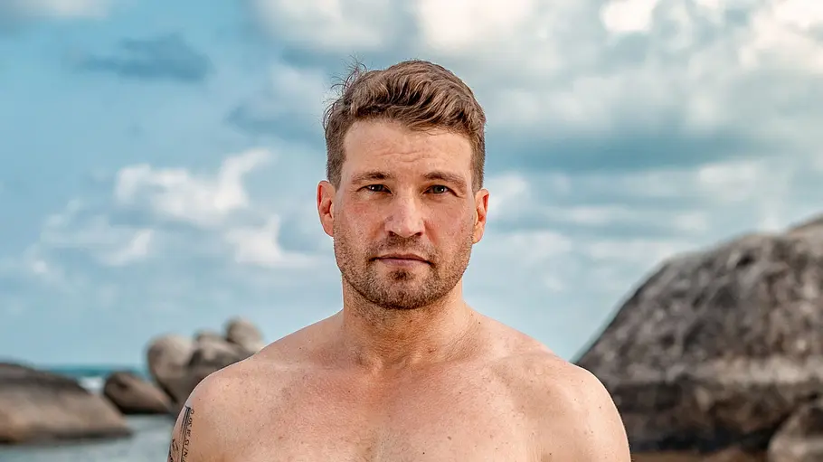 Raul Richter Ein muskulöser Mann steht oberkörperfrei am Strand und blickt direkt in die Kamera. Hinter ihm sind Meer, Felsen und ein bewölkter Himmel zu sehen, während er lässig die Hände in den Taschen seiner Badehose hat.