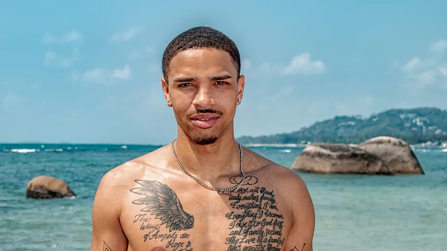 Robin Njie Ein junger Mann steht oberkörperfrei im flachen Meer und blickt in die Kamera. Er trägt eine helle Badehose, hat mehrere Tattoos auf Brust und Armen und wird von einer Küstenlandschaft mit Felsen und blauem Himmel im Hintergrund eingerahmt.