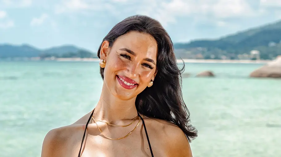 Zoe Müllner Eine Frau im Bikini steht lächelnd am Strand und blickt in die Kamera. Sie trägt Schmuck und hat lange dunkle Haare, während im Hintergrund das Meer, Felsen und eine hügelige Küstenlandschaft zu sehen sind.