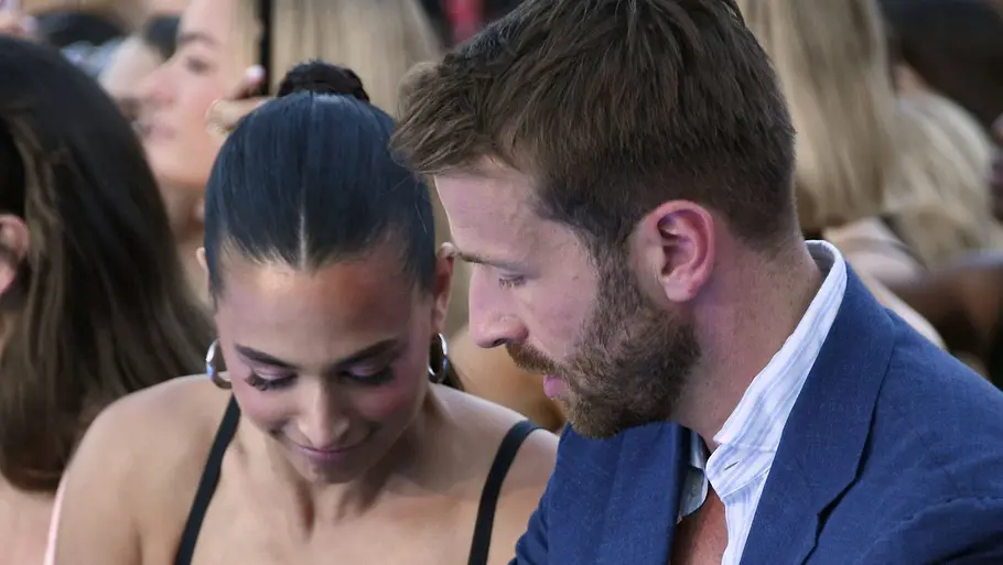 Amira Aly im schwarzen Kleid und Christian Düren im blauen Anzug sitzen zwischen anderen Gästen und blicken nach unten.