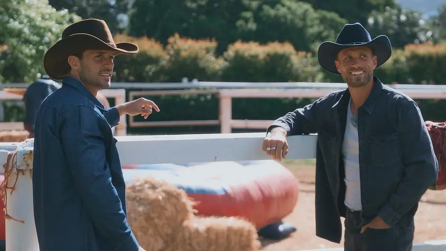 Bachelors Zwei Männer mit Cowboyhüten stehen an einem Weidezaun, lächeln und unterhalten sich auf einer Ranch. Im Hintergrund sind Strohballen und ein Rodeo-Hindernis zu sehen.