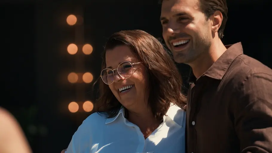 Bachelors Ein Mann und eine Frau stehen eng beieinander, lächeln in die Kamera und posieren für ein gemeinsames Foto. Der Mann hält ein Glas in der Hand, im Hintergrund sind unscharfe Lichter zu sehen.