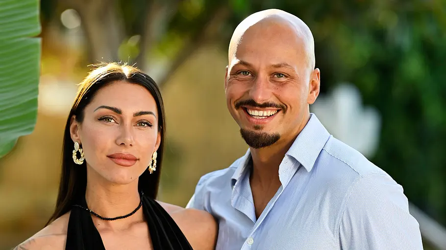 Porträt eines Paares im Freien vor einem unscharfen Hintergrund aus Bäumen und Pflanzen. Die Frau trägt ein schwarzes, figurbetontes Kleid mit tiefem Ausschnitt, auffällige Ohrringe und hat Tattoos auf einem Arm. Der Mann steht lächelnd neben ihr, trägt ein hellblaues Hemd und eine weiße Hose und hat einen kurz geschnittenen Bart. Beide blicken direkt in die Kamera und posieren eng beieinander in sommerlicher Umgebung.