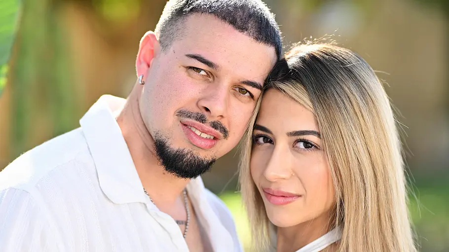 Porträt eines jungen Paares im Freien vor einem unscharfen Hintergrund aus Bäumen und Pflanzen. Der Mann trägt ein weißes Kurzarmhemd mit schwarzem Muster, hat einen kurzen Bart und legt einen Arm um die Taille der Frau. Die Frau trägt ein weißes Outfit mit bauchfreiem Oberteil und langem Rock oder Kleid, ihre langen blonden Haare fallen über die Schultern. Beide stehen eng beieinander, lehnen die Köpfe leicht aneinander und blicken direkt in die Kamera. Die Aufnahme vermittelt eine romantische und sommerliche Atmosphäre.