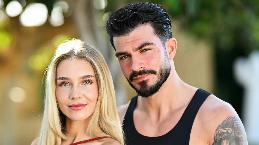 Michelle und Maki Reality-TV-Paar Michelle und Maki posiert eng umschlungen im Freien. Michelle trägt ein bordeauxrotes Kleid, Maki ein schwarzes Tanktop und helle Hose. Beide blicken lächelnd in die Kamera, während im Hintergrund Palmen und üppige Vegetation zu sehen sind.