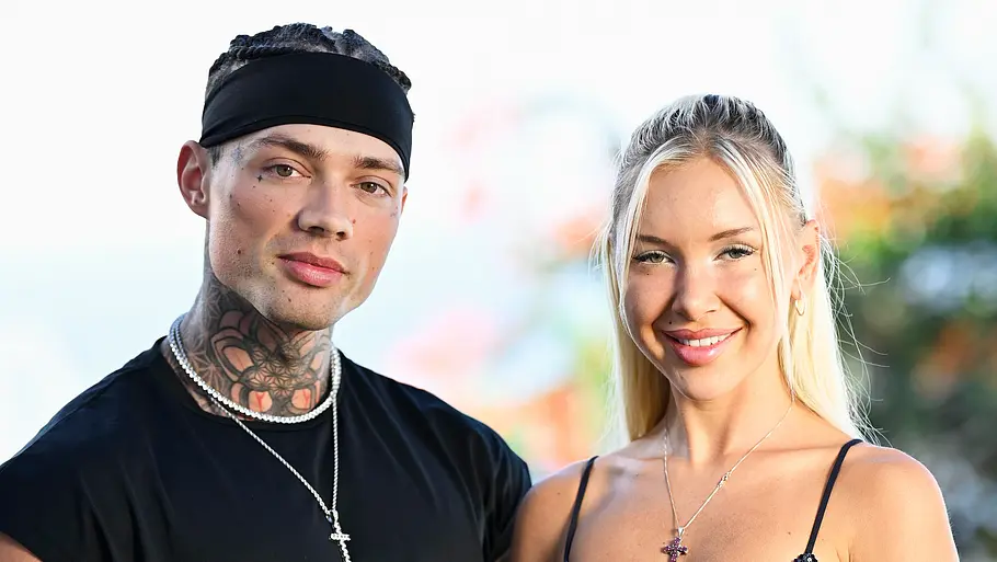 Josua Ein Mann mit Tattoos und schwarzem Outfit sowie eine blonde Frau in einem dunkelblauen Glitzerkleid stehen nebeneinander und lächeln für ein gemeinsames Porträt im Freien.