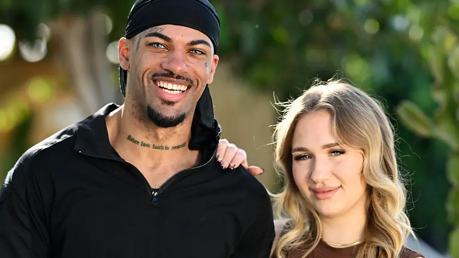 Porträt eines jungen Paares im Freien vor einem unscharfen Hintergrund aus Bäumen und Pflanzen. Der Mann trägt ein schwarzes Langarmshirt, graue Cargohose und ein schwarzes Kopftuch. Er lächelt breit in die Kamera und hat die Hände in den Hosentaschen. Die Frau steht eng an seiner Seite, legt eine Hand auf seine Schulter und trägt ein braunes, figurbetontes zweiteiliges Outfit. Beide blicken in die Kamera und posieren in einer entspannten, sommerlichen Umgebung.
