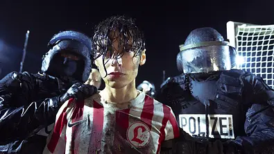 Eren M. Güvercin wird von zwei Polizisten abgeführt. Er trägt ein rot-weißes Trikot und ist auf einem Fußballplatz. - Foto: HBO