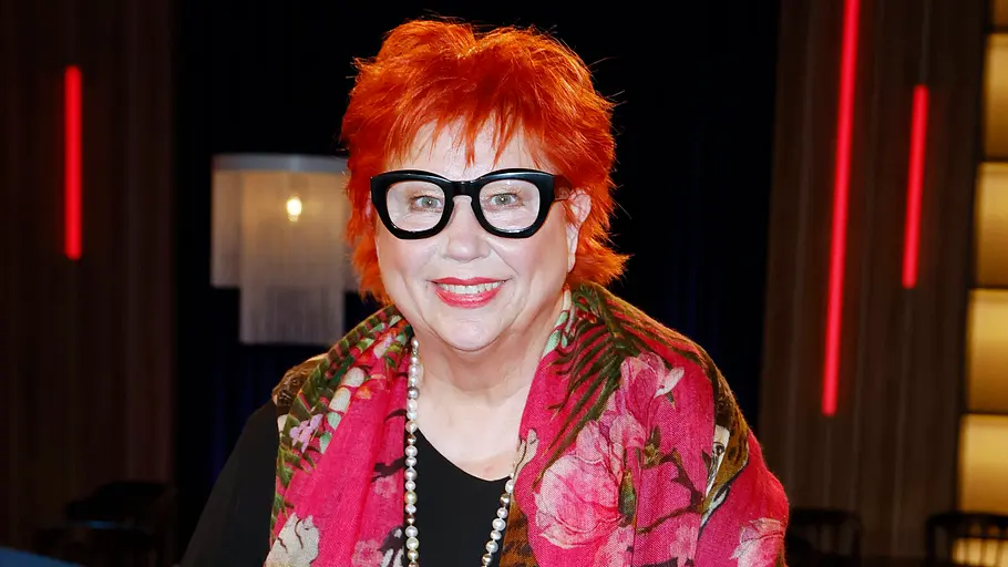 Eine lächelnde Frau mit kurzen roten Haaren und schwarzer Brille trägt einen pinken Blumenschal und eine lange Perlenkette.