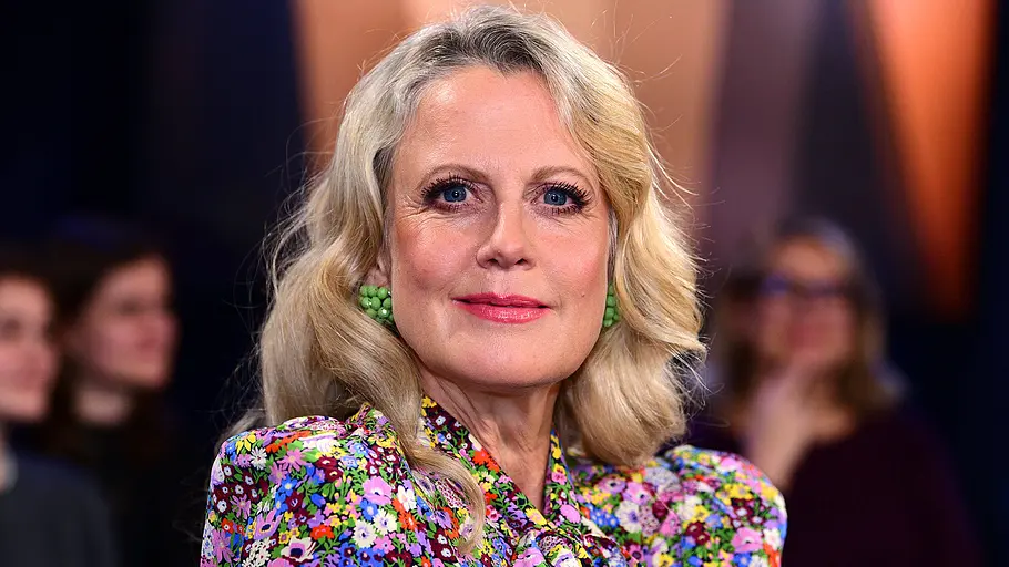 Barbara Schöneberger Barbara mit blonden Haaren und buntem geblümtem Kleid sitzt in einer Talkshow und blickt in die Kamera.