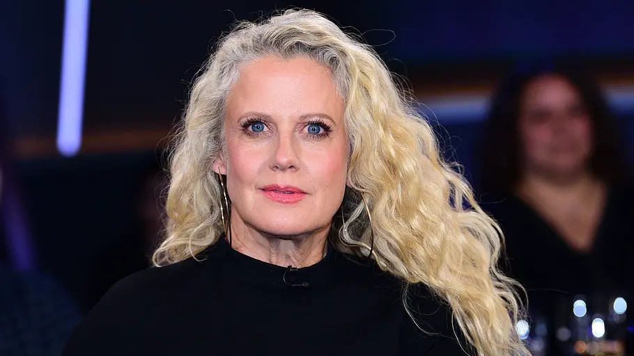 Barbara Schöneberger Lets Dance Barbara Schöneberger trägt ein schwarzes Oberteil und lange blonde Locken. Hinter ihr sitzen unscharf erkennbare Personen.
