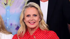 Eine blonde Frau mit offenen Haaren und auffälligem Make-up trägt eine rote Bluse mit weißen Punkten und Schleife am Kragen und lächelt direkt in die Kamera, während im Hintergrund weitere Personen zu sehen sind. - Foto: IMAGO / APress