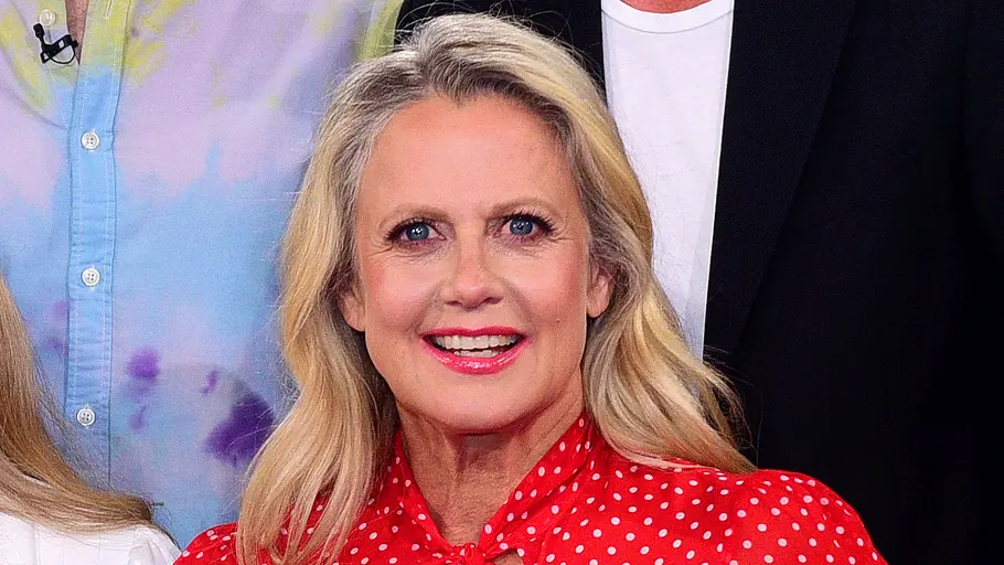 Barbara Schöneberger Eine blonde Frau mit offenen Haaren und auffälligem Make-up trägt eine rote Bluse mit weißen Punkten und Schleife am Kragen und lächelt direkt in die Kamera, während im Hintergrund weitere Personen zu sehen sind.