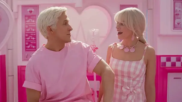Ken und Barbie stehen in einer knallpink eingerichteten, künstlich wirkenden Umgebung. Ken trägt ein rosa T-Shirt, Barbie ein rosa-weiß kariertes Kleid und eine auffällige pinke Halskette. Beide schauen sich an und wirken fröhlich überrascht.  - Foto: IMAGO / Bestimage