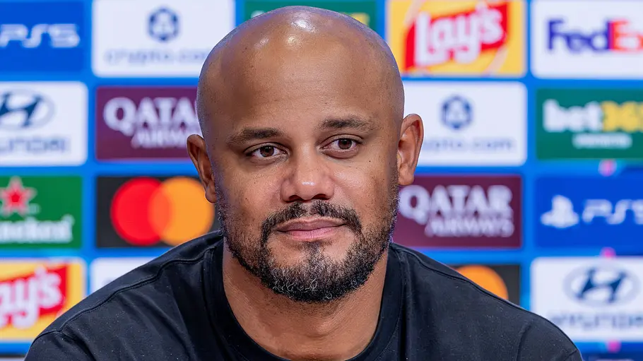 Bayern-Trainer Vincent Kompany im Krankenhaus. Vincent Kompany mit Glatze und kurzem Bart trägt ein schwarzes Shirt und sitzt vor einer blauen Wand mit Sponsorenlogos.