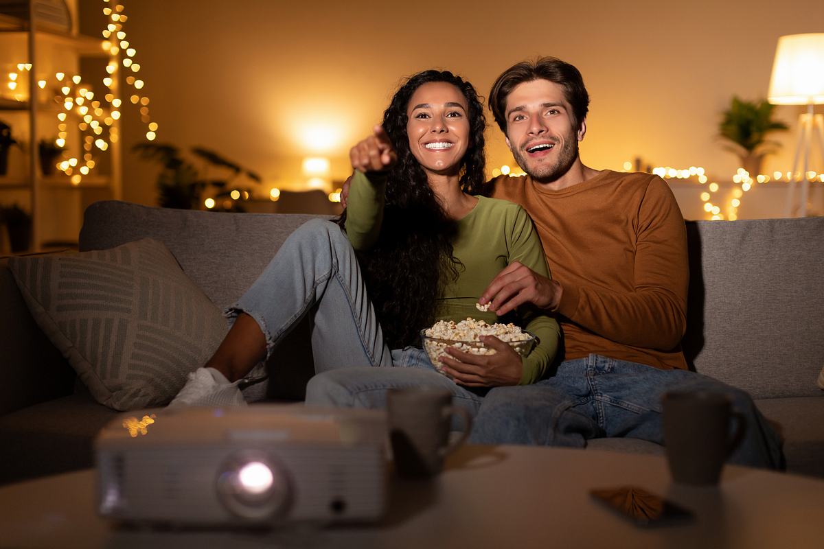 Beamer Angebot Ein Paar sitzt auf dem Sofa und schaut einen FIlm, sie haben eine Schüssel Popcorn bei sich. Vor ihnen steht ein Beamer.