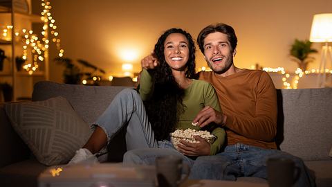 Ein Paar sitzt auf dem Sofa und schaut einen FIlm, sie haben eine Schüssel Popcorn bei sich. Vor ihnen steht ein Beamer. - Foto: istock/Prostock-Studio