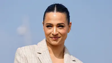 Amira Aly trägt einen hellen Blazer und die dunklen Haare zurückgebunden. Sie lächelt vor einem hellblauen Hintergrund. - Foto: IMAGO / Panama Pictures Amira Aly trägt einen hellen Blazer und die dunklen Haare zurückgebunden. Sie lächelt vor einem hellblauen Hintergrund. - Foto: IMAGO / Panama Pictures
