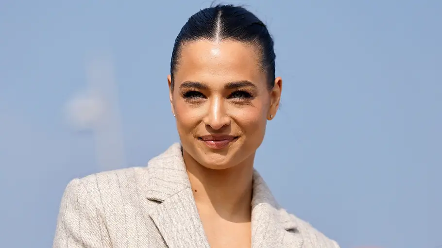 Bei Amira Aly und Christian Düren ist die Freude groß. Amira Aly trägt einen hellen Blazer und die dunklen Haare zurückgebunden. Sie lächelt vor einem hellblauen Hintergrund.
