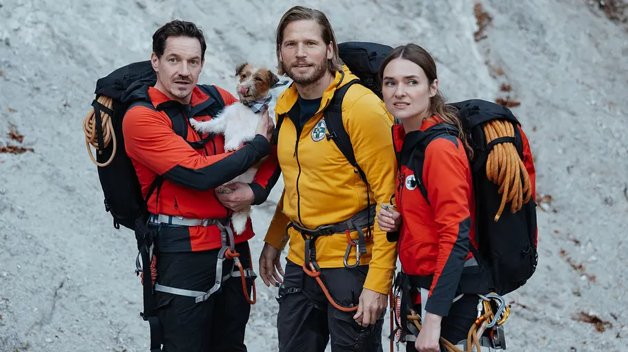Bei den „Bergrettern“ wird Staffel 18 auch abseits der gefährlichen Einsätze zur Zerreißprobe: Besonders Katharina und Markus stehen vor einer schweren Zeit. Tobi, Markus und Nina stehen mit Rucksäcken und Kletterausrüstung vor einer Felswand. Einer hält einen kleinen Hund im Arm.