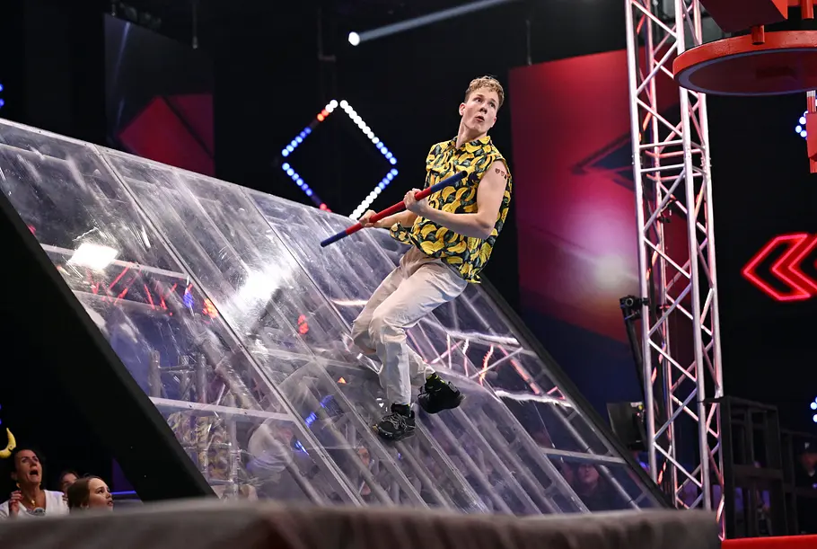 Ben Keilich ist Teilnehmer bei Ninja Warrior Germany 2026 Ben Keilich hält eine rot-blaue Stange vor einer schrägen transparenten Wand. Er trägt ein Bananenhemd und eine helle Hose.