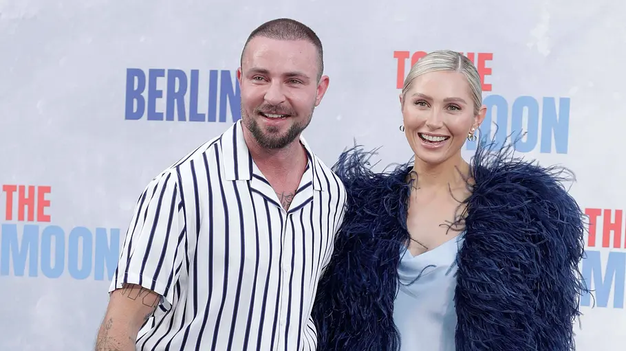 Benjamin Melzer und Sissi Hofbauer Benjamin Melzer im Streifenhemd und Sissi Hofbauer im hellblauen Kleid mit dunkler Federjacke posieren auf blauem Teppich.