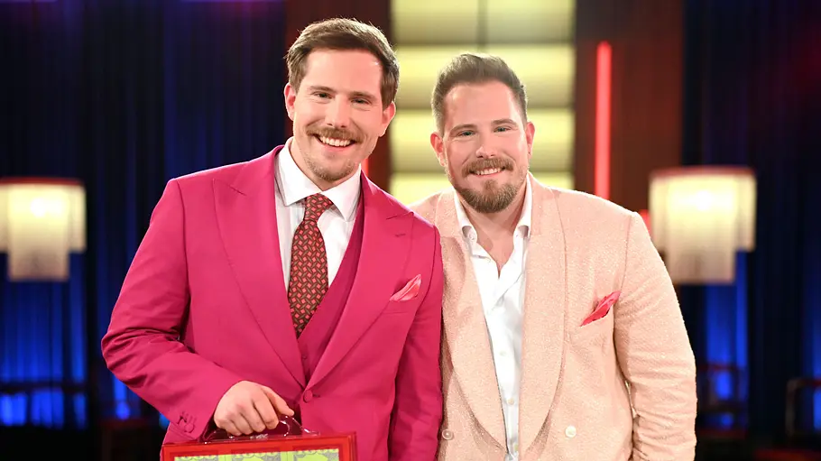 Dennis und Benni Wolter stehen lächelnd in pinkem und roséfarbenem Anzug nebeneinander. Einer hält einen roten Koffer.