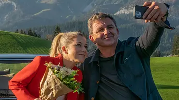 Das Auto von Martin Gruber steht vor einer Bergkulisse. Davor macht Martin Gruber (Hans Sigl) mit seinem Handy ein Selfie von sich und Karin Bachmeier (Hilde Dalik), die einen Blumenstrauß in der Hand hält.  - Foto: ZDF/Erika Hauri