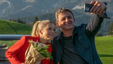 Das Auto von Martin Gruber steht vor einer Bergkulisse. Davor macht Martin Gruber (Hans Sigl) mit seinem Handy ein Selfie von sich und Karin Bachmeier (Hilde Dalik), die einen Blumenstrauß in der Hand hält. - Foto: ZDF/Erika Hauri Das Auto von Martin Gruber steht vor einer Bergkulisse. Davor macht Martin Gruber (Hans Sigl) mit seinem Handy ein Selfie von sich und Karin Bachmeier (Hilde Dalik), die einen Blumenstrauß in der Hand hält. - Foto: ZDF/Erika Hauri
