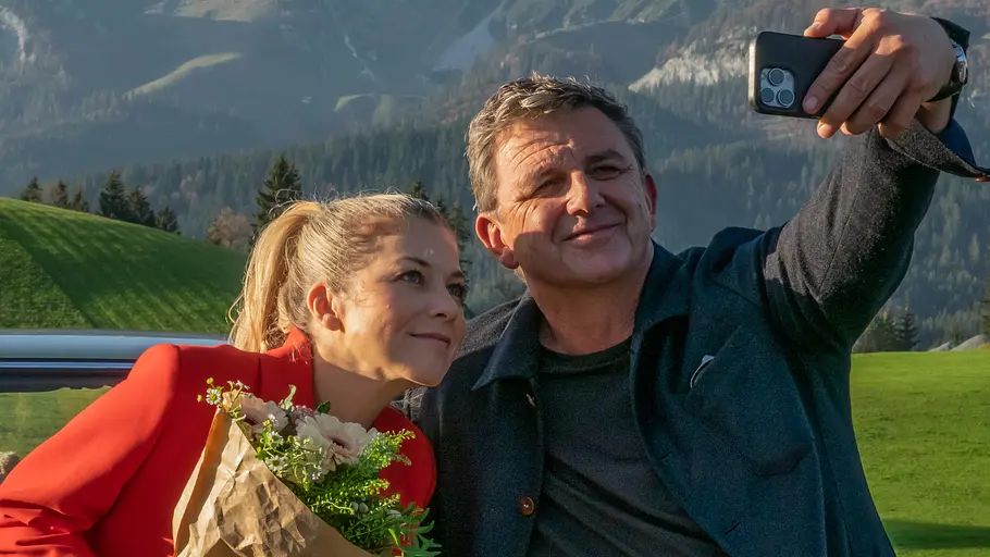 Hans Sigl ist steht seit Beginn an als Martin Gruber vor der Kamera. Das Auto von Martin Gruber steht vor einer Bergkulisse. Davor macht Martin Gruber (Hans Sigl) mit seinem Handy ein Selfie von sich und Karin Bachmeier (Hilde Dalik), die einen Blumenstrauß in der Hand hält.