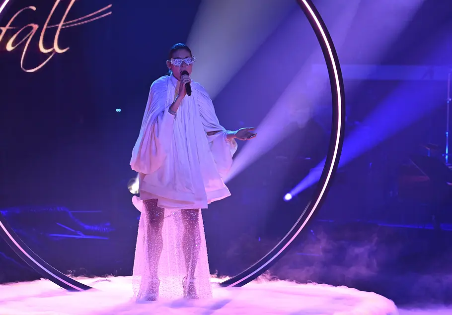 Bernarda Brunovic Halbfinale The Voice 2025 Bernarda Brunovic trägt ein weißes Kleid und eine glitzerne Sonnenbrille. Auf der Bühne steht ein großer leuchtender Kreis und Nebel.