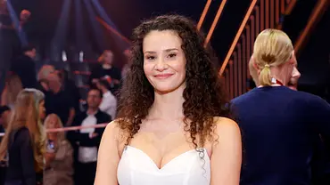 Betty Taube lächelt im weißen Kleid bei einer Veranstaltung von „Let’s Dance“, im Hintergrund sind weitere Personen und Lichter der Show zu sehen. - Foto: IMAGO / Panama Pictures