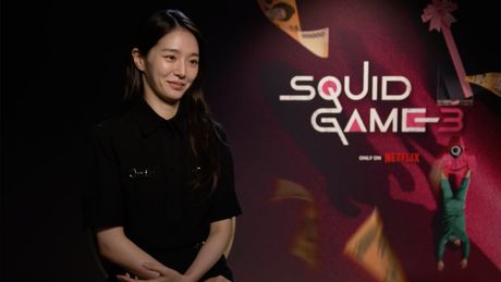 Park Gyu-young sitzt vor einem „Squid Game“-Poster und lächelt. - Foto: Netflix