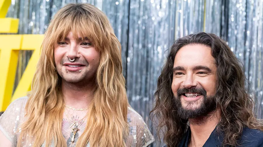 „Kaulitz & Kaulitz“: Staffel 3 die Brüder auf dem roten Teppoich Bill Kaulitz mit blonden Haaren mit Piercings und durchsichtigem, glitzerndem Oberteil und Tom Kaulitz, dunkelhaarig mit Bart im dunklen Sakko vor einem silbern schimmernden Vorhang bei der Premiere von der 3. Staffel der Netflix-Serie „Kaulitz & Kaulitz“