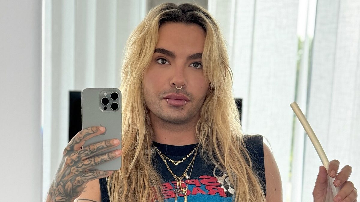 Bill Kaulitz Bill Kaulitz posiert vor dem Spiegel. Man sieht seine tätowierte Hand, in der er sein Handy hält, und er schaut direkt in die Kamera. In der anderen Hand hält er ein Getränk.