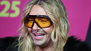 Bill Kaulitz lacht bei einem Event vor pinkem Hintergrund und trägt lange blonde Haare, große getönte Sonnenbrille und mehrere Piercings. - Foto: IMAGO / dts Nachrichtenagentur