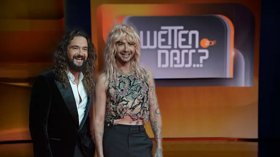 Bill und Tom Kaulitz „Wetten, dass..?“ Ticket-Andrang. Bill und Tom Kaulitz posieren lächelnd vor dem „Wetten, dass..?“-Logo, einer im Anzug, einer im verzierten bauchfreien Top.