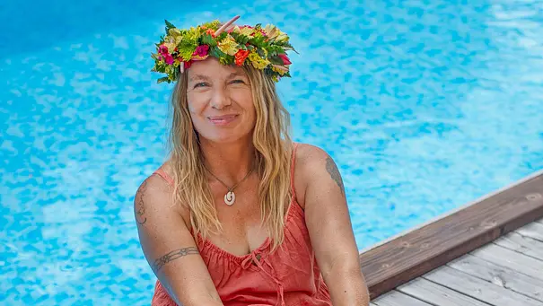 Manu Reimann trägt einen Blumenkranz auf dem Kopf und sitzt lächelnd vor einem Pool. - Foto: Kabel Eins / Lucie XYZ
