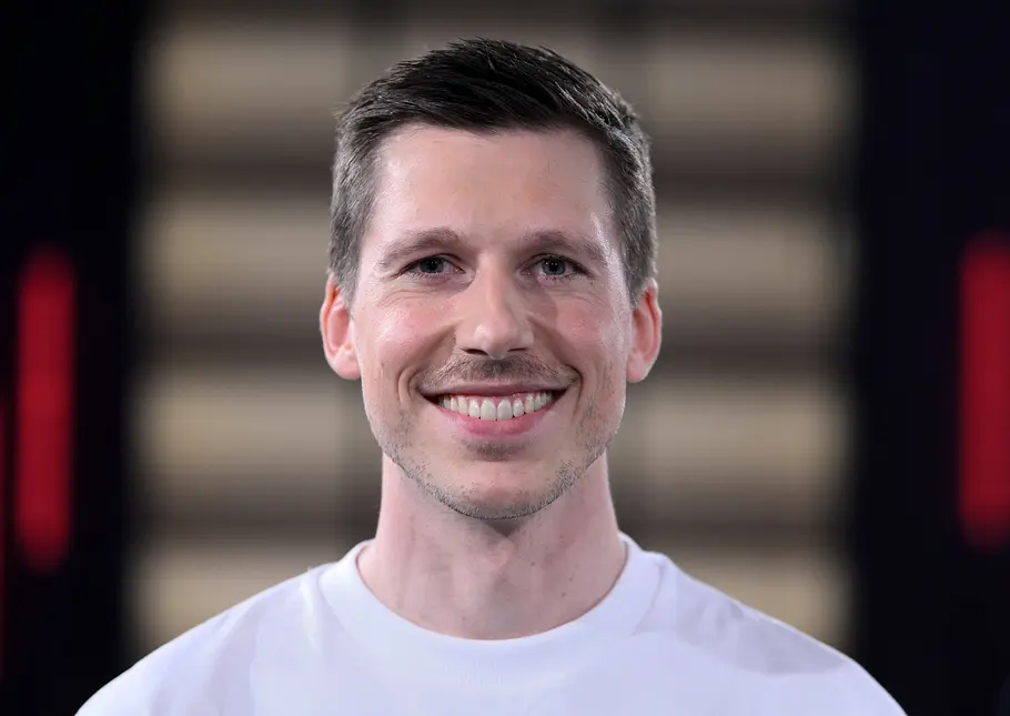 Björn von Morgenstern trägt ein weißes T-Shirt und lächelt freundlich in die Kamera. Der Hintergrund ist unscharf und dezent beleuchtet.