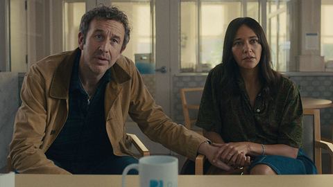 Chris ODowd und Rashida Jones aus der 1. Episode der 7. Staffel von Black Mirror. Beide sitzen auf Stühlen vor einem Tisch, sie sehen überrascht aus. - Foto: Netflix